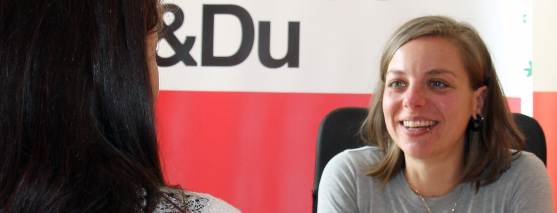 Caritas Beratungsgespräch – Unterstützung und Hilfe Eine Caritas-Mitarbeiterin führt ein freundliches Beratungsgespräch mit einer Frau. Im Hintergrund ist ein Caritas & Du-Banner zu sehen.