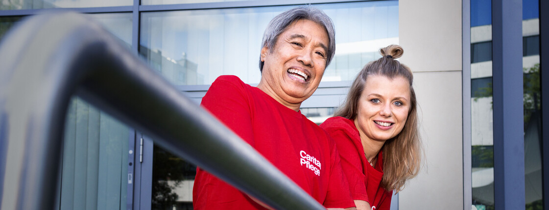 Caritas Pflege_Header_2023 lachender Mann und lachende Frau mit roten Caritas T-Shirts stehen hinter Geländer und schauen in Kamera