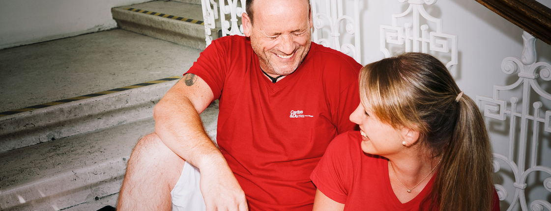 Ein Mann und eine Frau mit roten T-Shirts mit Caritas-Logo sitzen auf den Stiegen und lachen gemeinsam. Die Frau hat einen Aktenordner in der Hand.